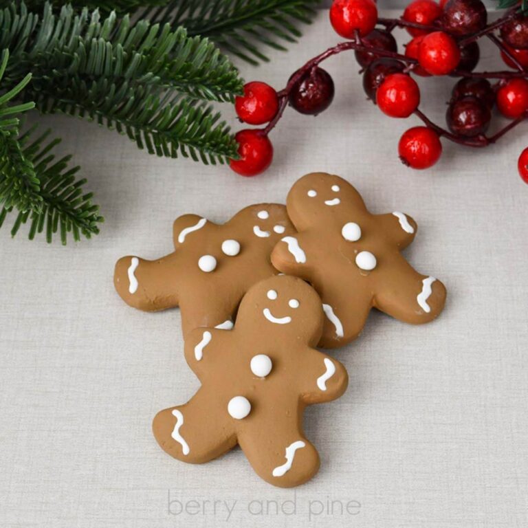 Square, close up image of three brown, fake gingerbread cookies with faux white icing on a linen background with pine and berries.